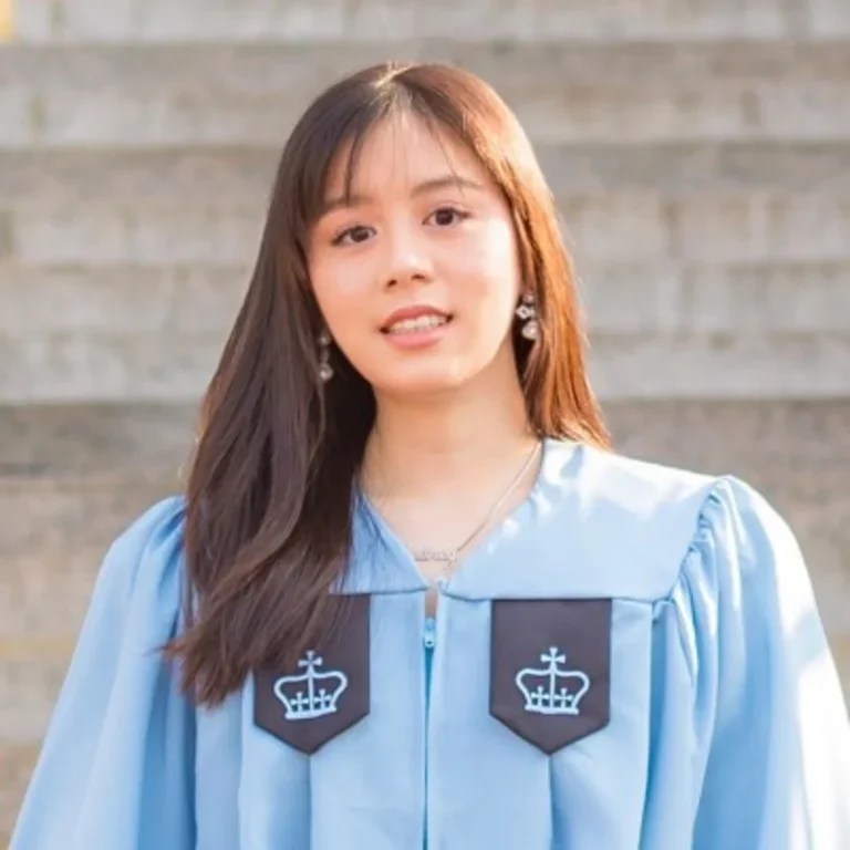 A professional headshot of Eugenie Wasito wearing light blue Columbia University graduation regalia, smiling in front of a blurred outdoor background.