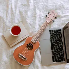 A stock image of a ukulele, tea and a laptop on a bed.