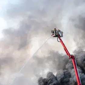 Firefighter on firetruck ladder using a hose with smoke building in the background