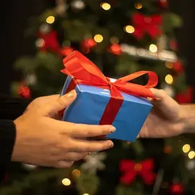 A photo shows a person handing a small gift to another person as they stand in front of a Christmas tree.