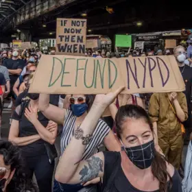 Protesters in face masks hold up signs that read "BLM" and "Defund NYPD."