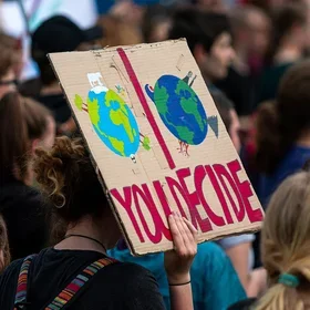 Sign at a environmental advocacy demonstration that reads "You Decide" with a picture of a healthy Earth with trees and animals side by side with a picture of a darker Earth with power plants, volcanoes, and tsunamis