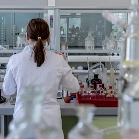 A scientist wearing a white lab coat is seen from behind as she works in a modern laboratory. The workspace is filled with various glassware, bottles, and laboratory equipment, including beakers, flasks, and pipettes. Shelves and counters are organized with chemical containers and instruments, reflecting a busy and well-equipped scientific lab.