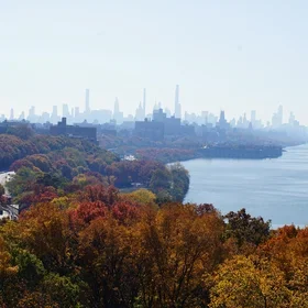 Autumn view of New York City skyline with the Hudson River, with orange, brown, and yellow trees in the foreground and modern skyscrapers rising in the background under a misty sky.