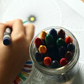 A child’s hand coloring in a coloring book with a box of multicolored crayons.