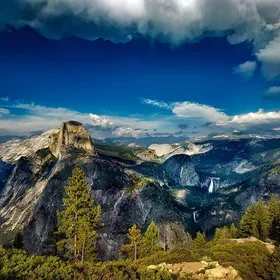 A rugged mountain ridge rises above a dark forest of evergreen trees, with wispy clouds drifting just below the peaks under a bright blue sky.