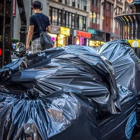 Piles of black garbage bags stacked beside a metal street trash can on a busy New York City sidewalk at dusk, with pedestrians walking past and storefront lights glowing in the background.