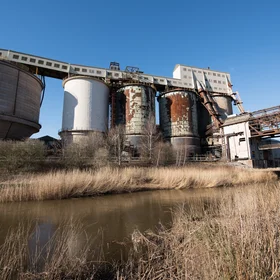 A chemical plant with multiple large cylindrical storage tanks, processing structures, and elevated conveyor systems stands beside a narrow waterway. Rust-streaked silos and industrial buildings dominate the site, while reeds and sparse vegetation line the canal in the foreground.