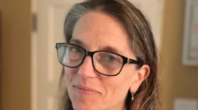 Profile of woman in a dark blazer with long dark blonde hair and thick dark glasses smiling