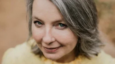 Above shot of smiling woman in light yellow blouse with delicately ruffled collar.  Shoulder length ashy blonde bob and simple pendant necklace