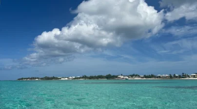Calm turquoise Caribbean water stretches toward a low-lying island shoreline beneath large white clouds and a bright blue sky.